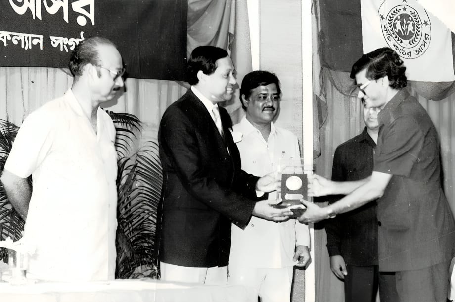 A black-and-white photograph: a man in a dark suit handing a mounted plaque to a young woman on a stage, with two men standing behind them
