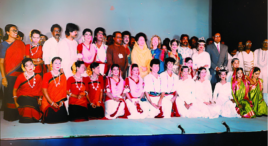 A wide row of children in costume seated on a stage with adult guests standing behind them