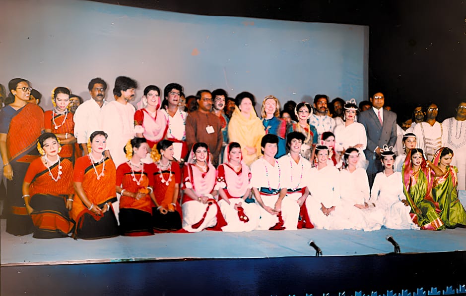 A colour photograph: two rows of performers in red, white and green costume seated on a stage, with a row of adults standing behind them