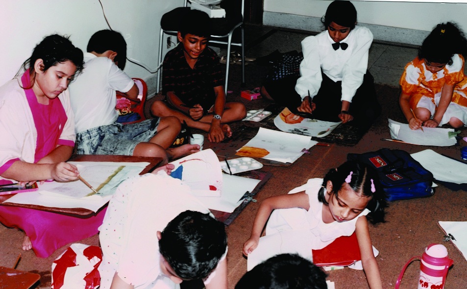 Children sitting on the floor of a hall, painting on large sheets of paper spread out in front of them. Children painting on the floor at a Kochi Kontho art session.